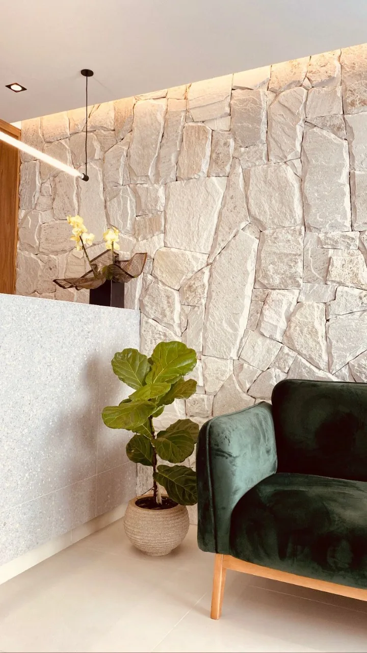 Ambiente interno com parede de pedra clara, poltrona verde e vegetação.